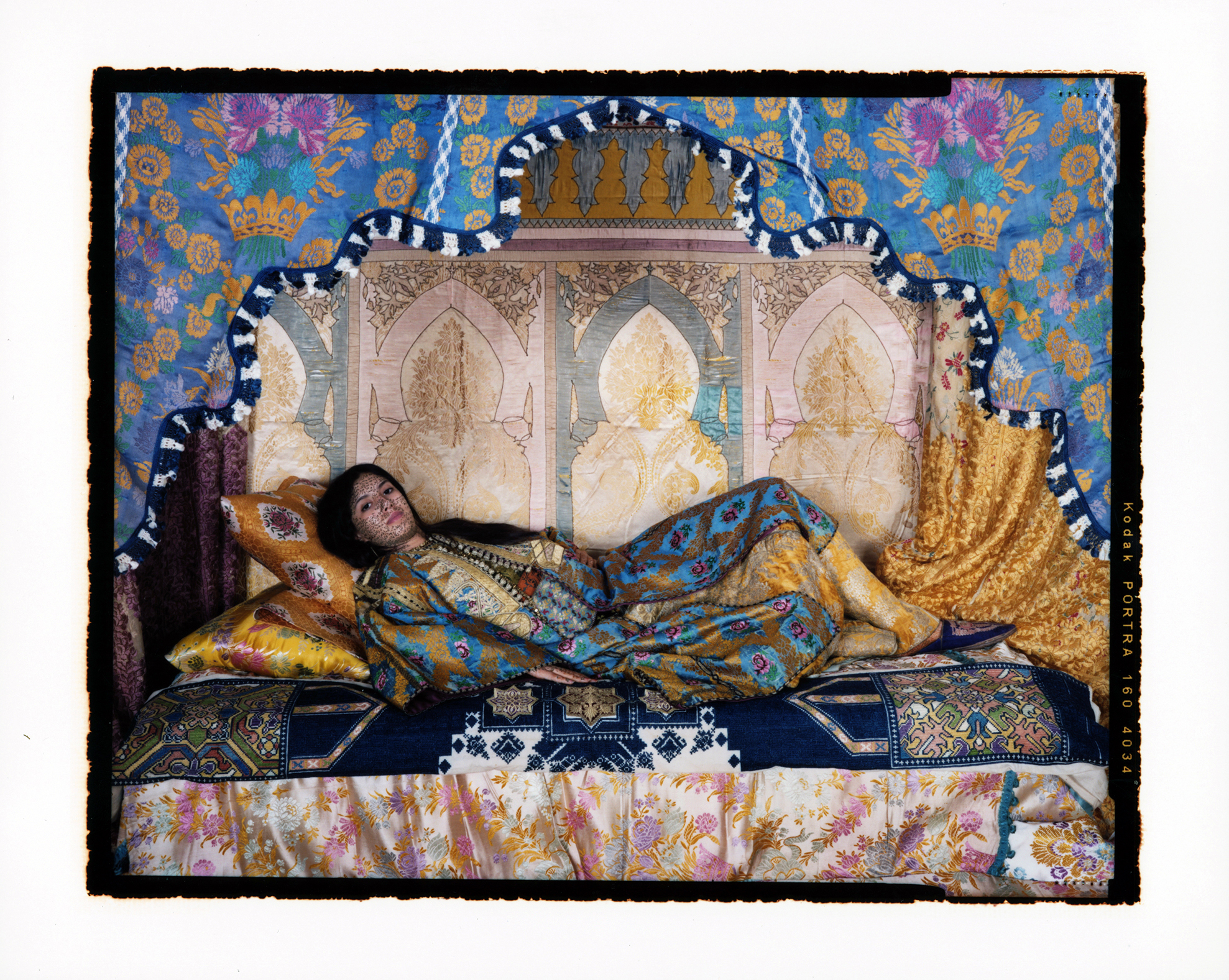 A colorful, staged photograph features a light-skinned woman with long dark hair reclined on a luxurious day bed covered in textiles featuring patterns from the Arab world. Her head is propped up on two, yellow patterned silk pillows and she looks straight at the camera. Her skin is marked with black writing, unreadable at the distance the phot is taken. She is draped in colorful, patterned robe that is different from, but in the same color scheme of, the patterns behind her on the wall and those on the bed.