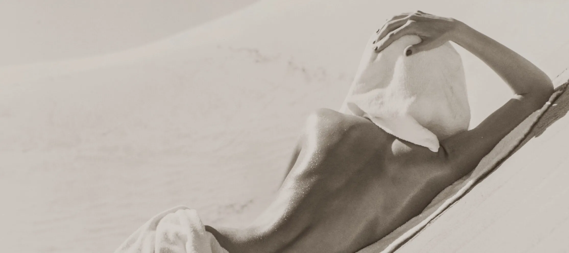 A black and white photograph of a light-skinned figure lying naked on a sand dune. The figure faces away from the camera with a white towel covering the back of their head and another towel draped across their hips, covering their bottom. Rolling sand dunes fill the background of the photograph.