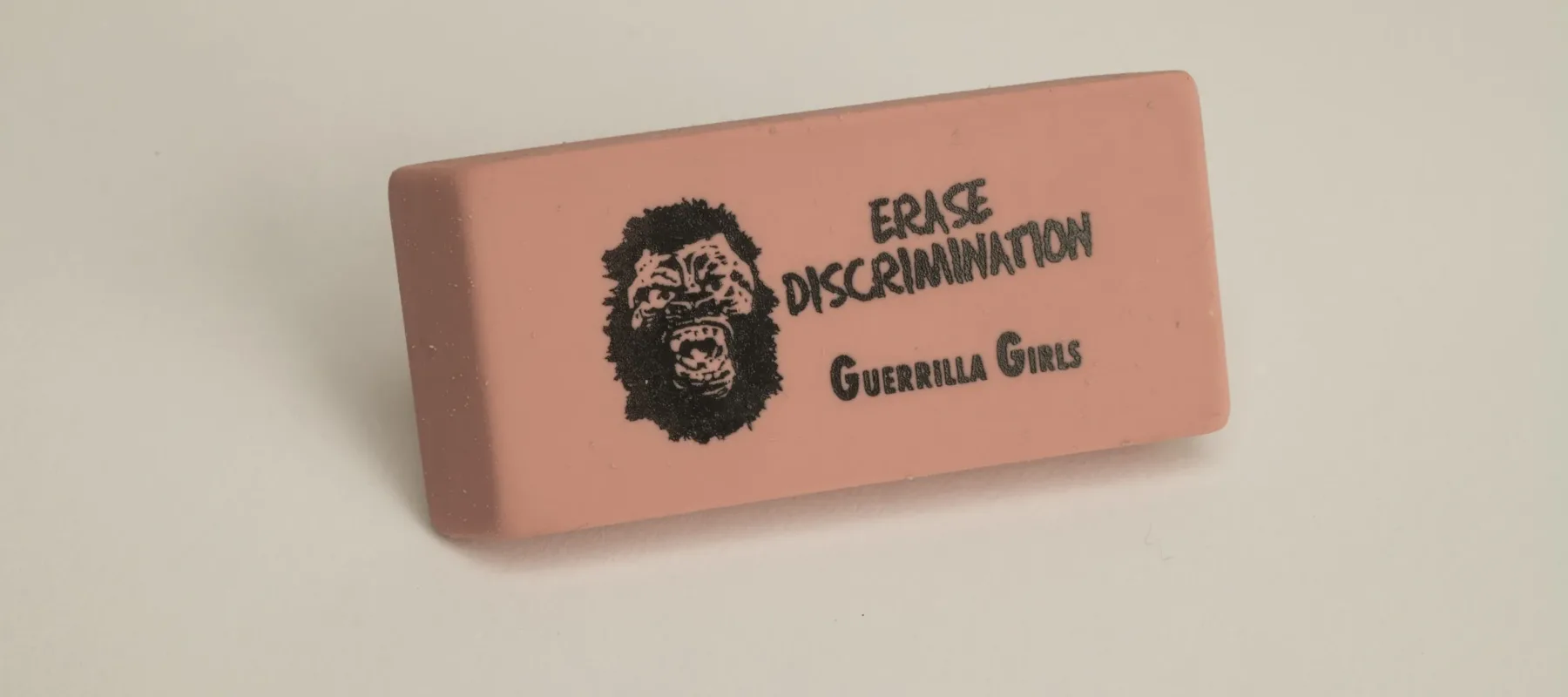 A rectangular pink eraser against a white background. Printed on the eraser in black is a yelling gorilla face to the left of the words, "ERASE DISCRIMINATION" above the words "Guerrilla Girls."