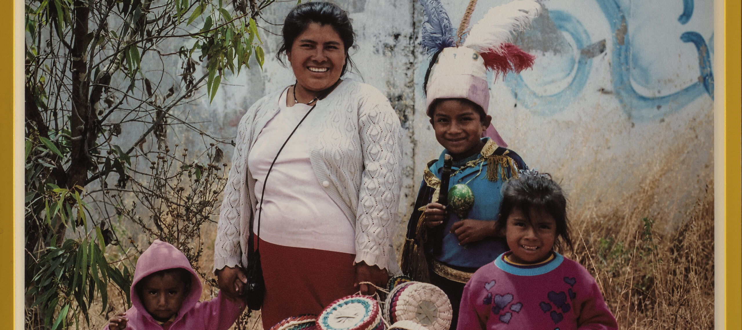 A yellow-framed photograph of four medium-skinned individuals—an adult woman and three young children. They stand in front of a cement wall with graffiti and hold multiple colorful baskets in their hands. Printed below them is the word 'Oaxaca'.