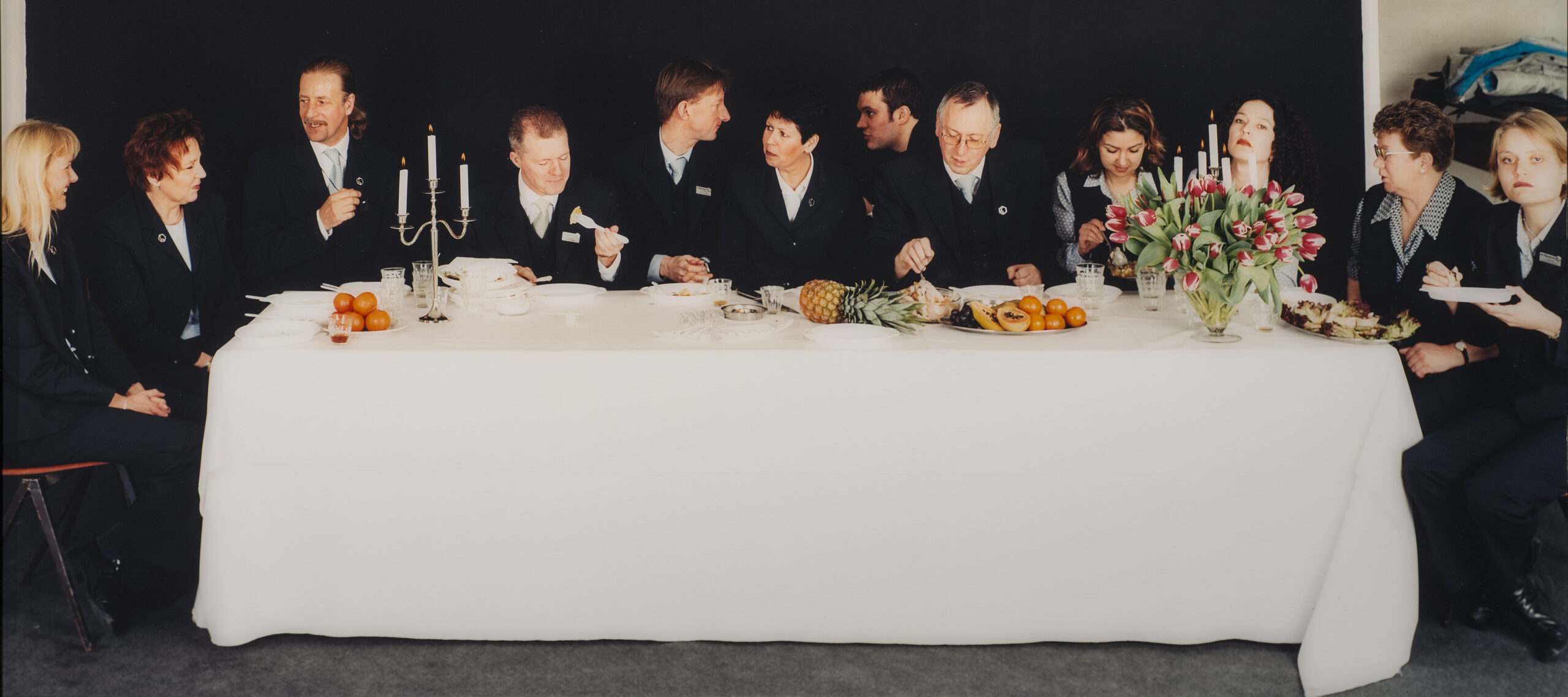 Twelve light-skinned men and women in dark suits sit on the ends and far side of rectangular table situated horizontally to the viewer. Covered with a white cloth, the table holds 2 candelabra, a flower bouquet, and assorted fruit. The people eat, chat, or look out at viewers.