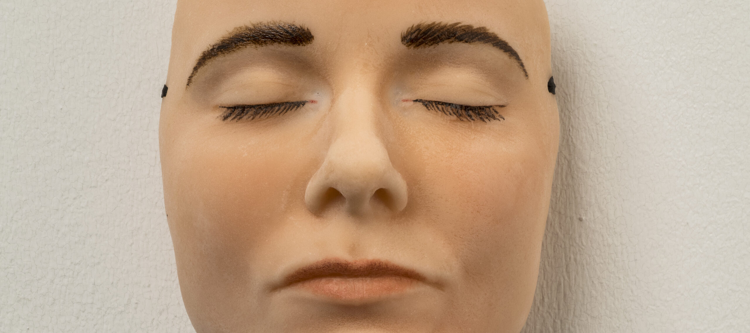 A highly realistic mask of a light-skinned face with dark brown eyebrows and eyelashes that appears to be sleeping—its eyes and mouth are closed and relaxed. The mask hangs on a white wall.