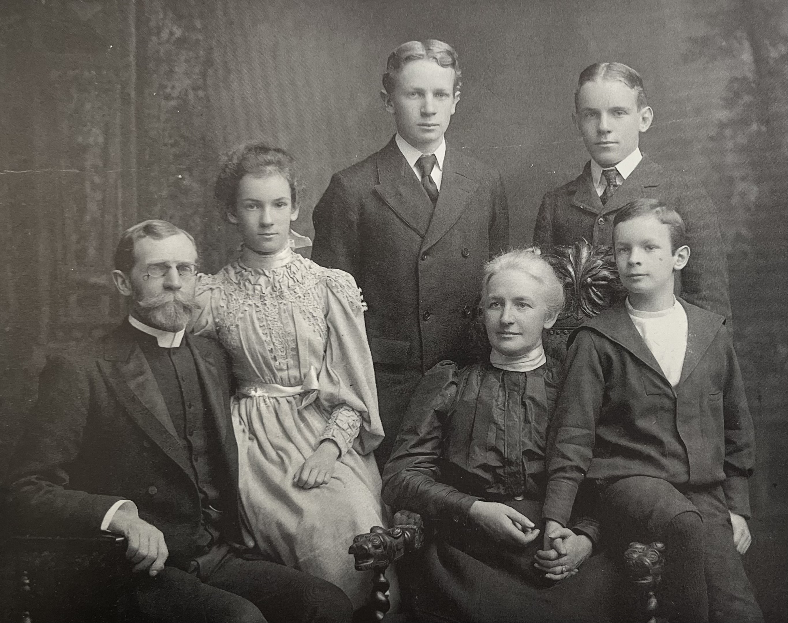 A black-and-white family portrait from the 1800s features six light-skinned individuals: two parents and their three sons and one daughter, who are pre-teen or teen age. Everyone wears dark dress except for the daughter, who is in a light, lace dress with a high collar. They all stare at the camera.