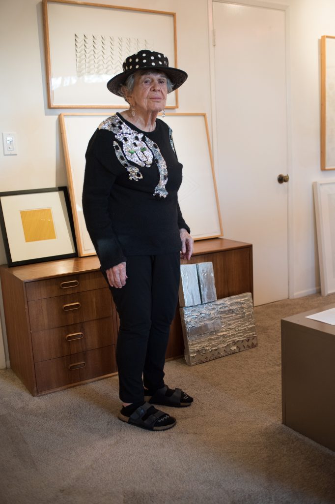 A woman with a light skin tone and short gray hair stands in her home surrounded by art pieces. She is wearing a funky black hat with polka dots, a black sweater with a sequin leopard, and black Birkenstock sandals.