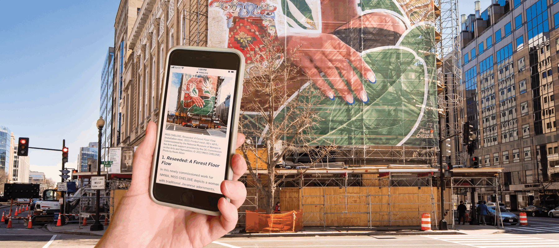 A light-skinned hand holds a mobile phone towards a four-story mural installed on the façade of the National Museum of Women in the Arts. The mural shows the upper half of a woman with a medium-light skin tone surrounded by various colorful plants and symbols. The phone’s screen faces the viewer and contains a small image of the mural and text.