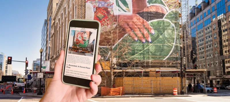 A light-skinned hand holds a mobile phone towards a four-story mural installed on the façade of the National Museum of Women in the Arts. The mural shows the upper half of a woman with a medium-light skin tone surrounded by various colorful plants and symbols. The phone’s screen faces the viewer and contains a small image of the mural and text.