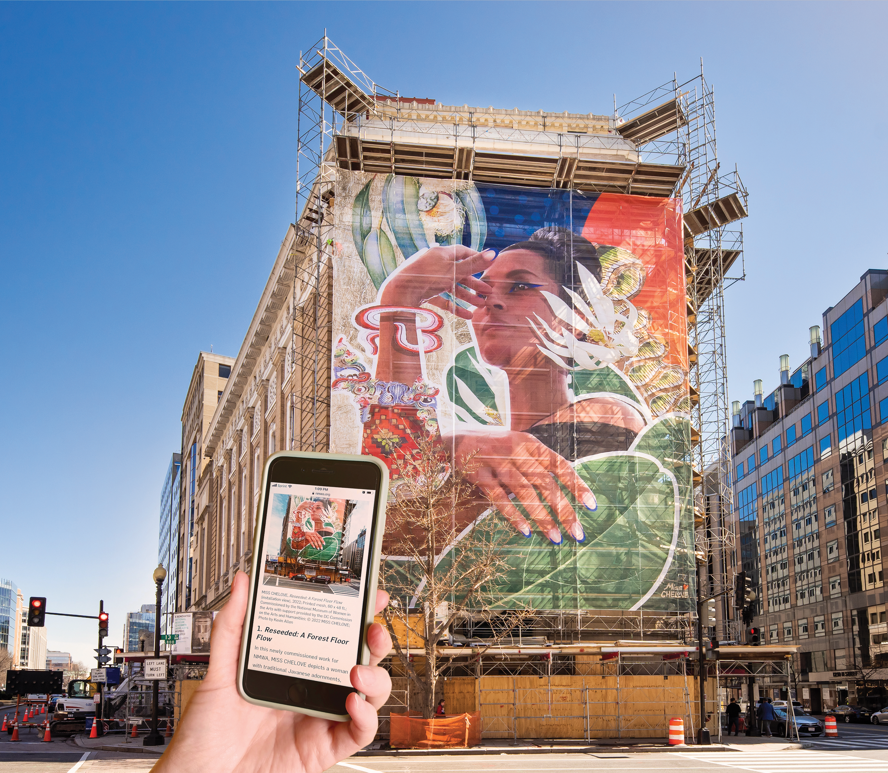 A light-skinned hand holds a mobile phone towards a four-story mural installed on the façade of the National Museum of Women in the Arts. The mural shows the upper half of a woman with a medium-light skin tone surrounded by various colorful plants and symbols. The phone’s screen faces the viewer and contains a small image of the mural and text.