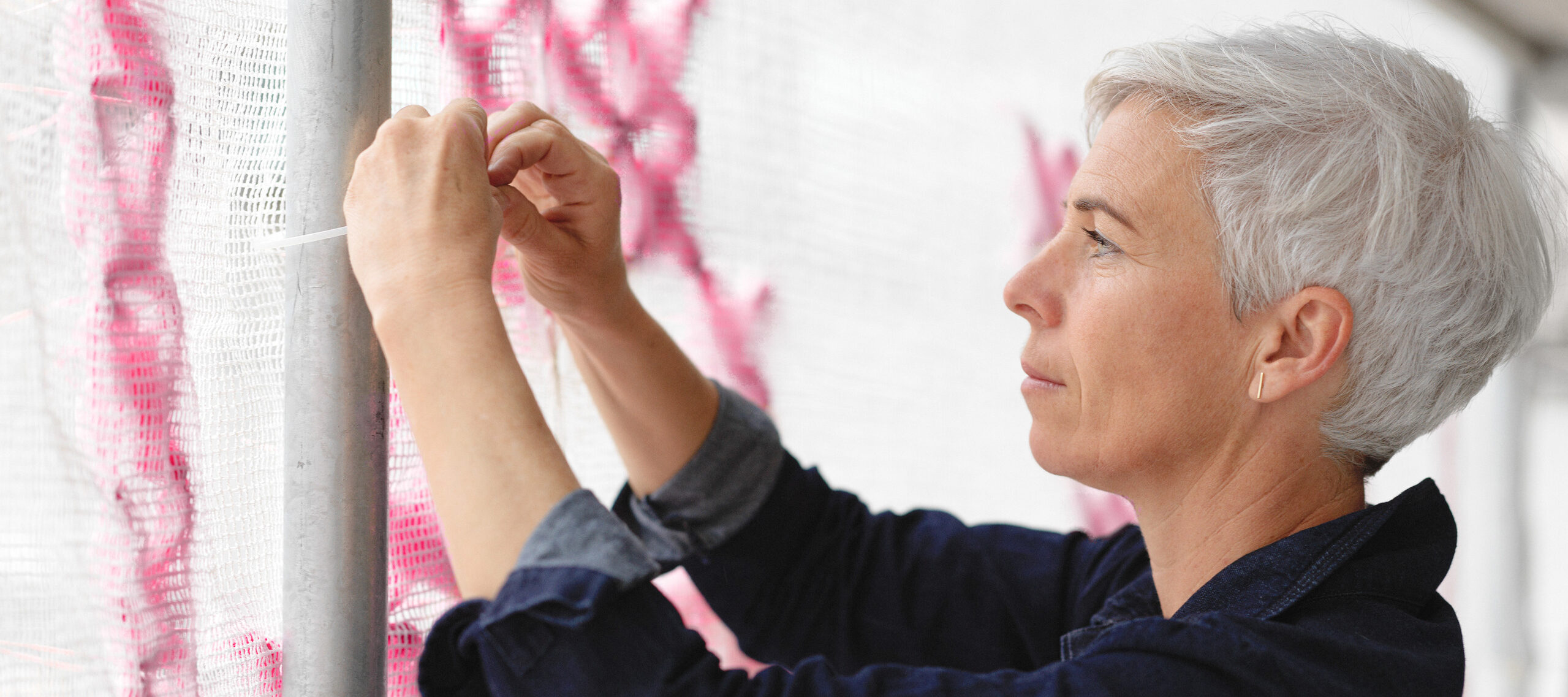 A light-skinned white woman with short white hair stands in profile, tying white scrim with bright pink stitching to a metal pole.