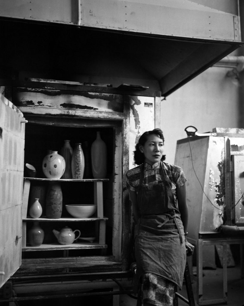 A black-and-white photograph shows an Asian American woman who wears an apron and leans on a chair in a ceramics workshop next to a furnace. The door to the furnace is open and offers a glimpse inside, where finished works, including vases and bowls, are placed. 