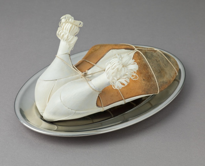 A photograph of what at first sight looks like a chicken with paper frills on a tray. On closer inspection, not at all: Two white high heels are tied together with a cord and intentionally arranged in a way to make them look like chicken legs.