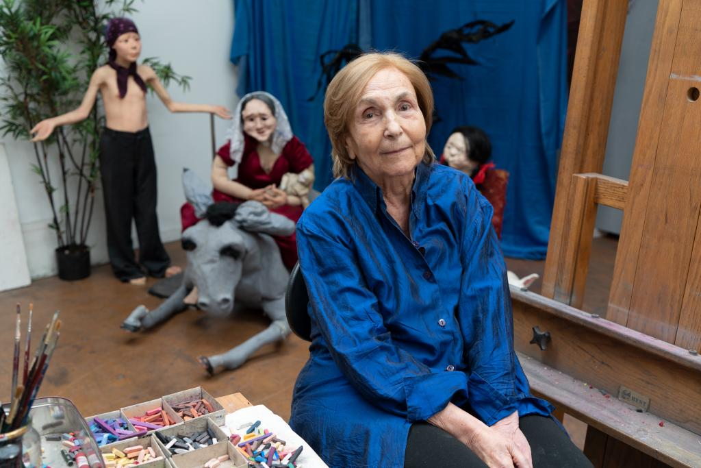 An older woman with dark blonde hair and a white skin tone sits in her art studio. She wears a blue shirt and sits next to a table with painting materials and an easel. Behind her are three life-size paper mache figures of a woman, a donkey, and two children.