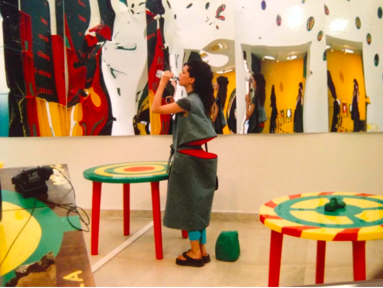 A woman in a costume stands in a room that looks like a funhouse, with colorful tables and a wall of distorting mirrors. The gray and red costume is divided into two parts and opens on her back, resembling an open mouth. 