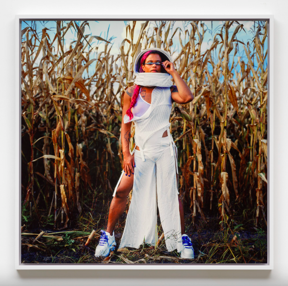 A photograph of a woman with a medium skin tone who wears an all-white outfit and stands before a cornfield. She wears a white astronaut’s helmet which gives the scene a futuristic quality.