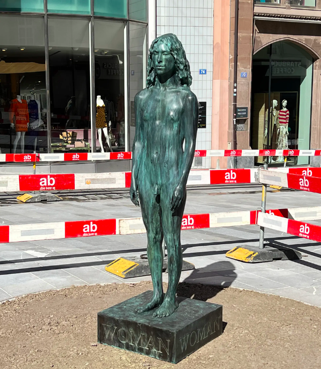 A sculpture of a trans woman in the style of ancient Greek statuary stands in the pedestrian zone of Basel. The sculpture is on a pedestal that says “Woman” and has a weathered look.