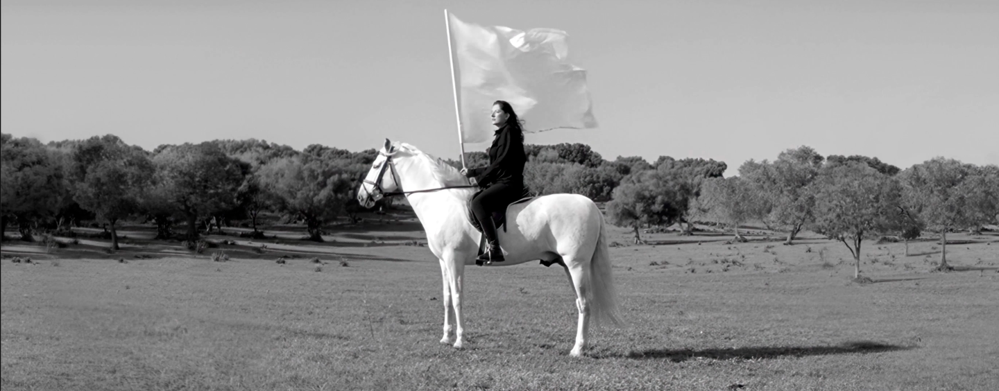  A black and white film still of a woman who sits on a white horse and holds a white flag. She is on a field surrounded by trees, and her all-black outfit contrasts the horse and flag.