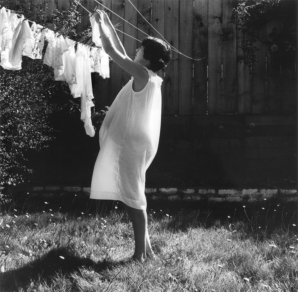 In this black-and-white photo, a light-skinned woman with short, dark hair stands in a backyard, hanging what looks to be baby clothes on a clothesline that is attached to w wooden fence. She wears a white sleeveless nightgown, under which the outline of her pregnant body is visible as the sun illuminates her and makes the gown transparent.