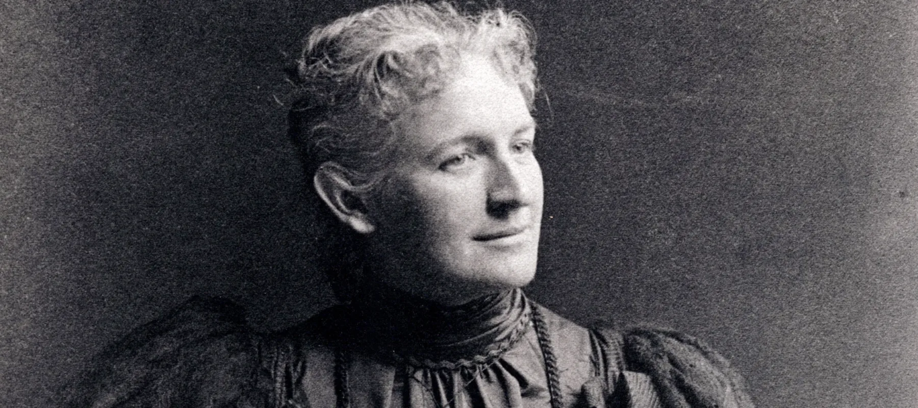 A black-and-white portrait photograph of a light-skinned woman from the late 1800s.She wears a dark, ruffled dress that has a high collar and puffy sleeves. Her light hair is pulled back and she looks to the right, slightly smiling, while one hand rests on her hip and the other in her lap, atop a small leather bag.