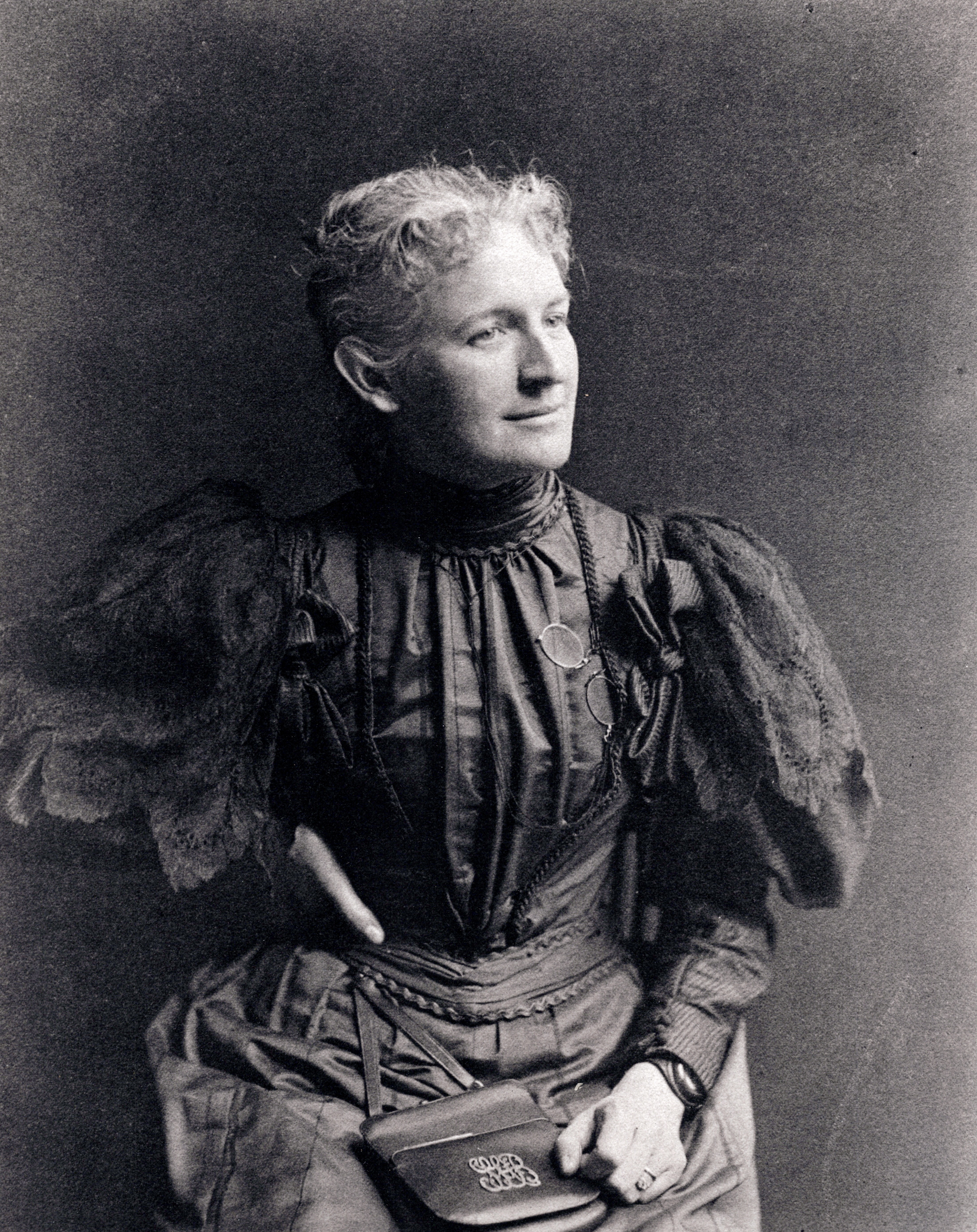 A black-and-white portrait photograph of a light-skinned woman from the late 1800s.She wears a dark, ruffled dress that has a high collar and puffy sleeves. Her light hair is pulled back and she looks to the right, slightly smiling, while one hand rests on her hip and the other in her lap, atop a small leather bag.