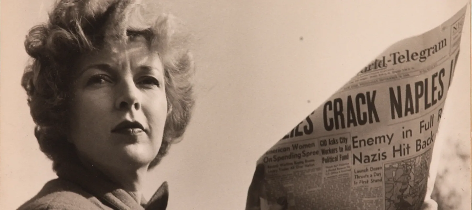 A black-and-white photograph of a light-skinned adult woman holding a newspaper with news about World War II. She wears a coat and her short, curly hair is caught in the wind.