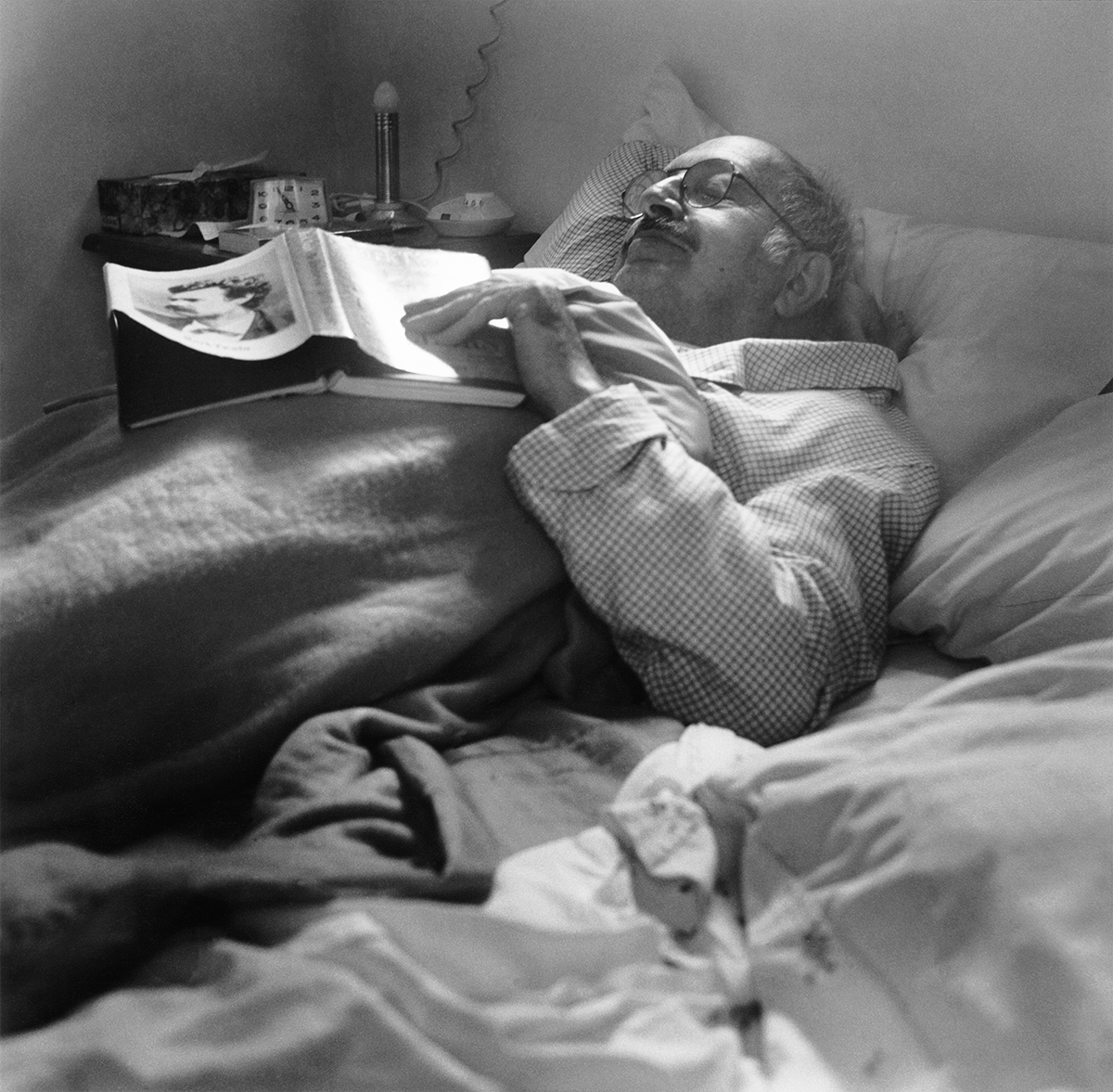 In this black-and-white photo, an older, light-skinned man wearing glasses lays in bed under the covers with his eyes closed as an open book rests on his chest. One hand holds the book and the other is reclined behind his head. A cluttered night stand is visible to the left. The man looks peaceful.