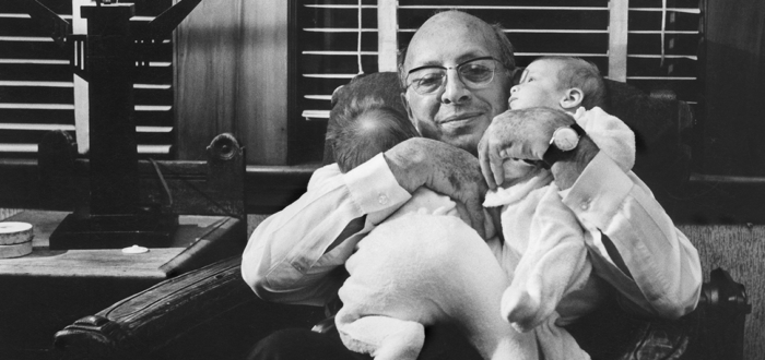 In this black-and-white photo, an older, light-skinned man wearing glasses sits in a dark armchair. He holds two babies on his chest, both of his arms cradling them close. Both babies wear white, footed onesies. One baby faces away from the camera on its stomach, while the other is cradled on its back at an angle, looking to the left. The man smiles directly at the camera.