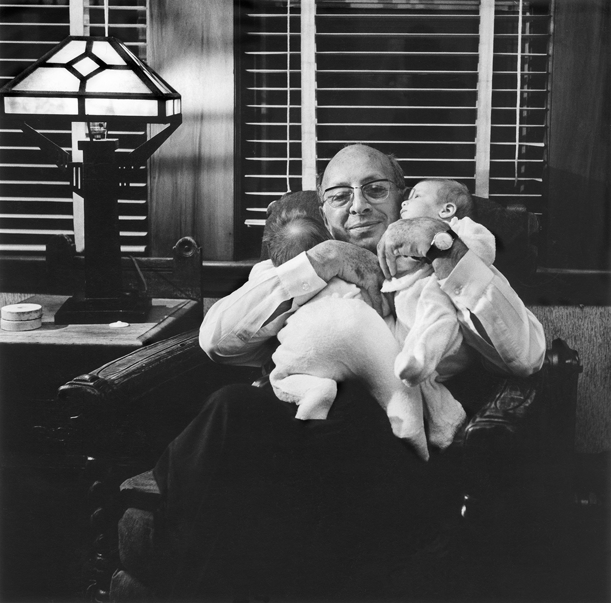 In this black-and-white photo, an older, light-skinned man wearing glasses sits in a dark armchair. He holds two babies on his chest, both of his arms cradling them close. Both babies wear white, footed onesies. One baby faces away from the camera on its stomach, while the other is cradled on its back at an angle, looking to the left. The man smiles directly at the camera.