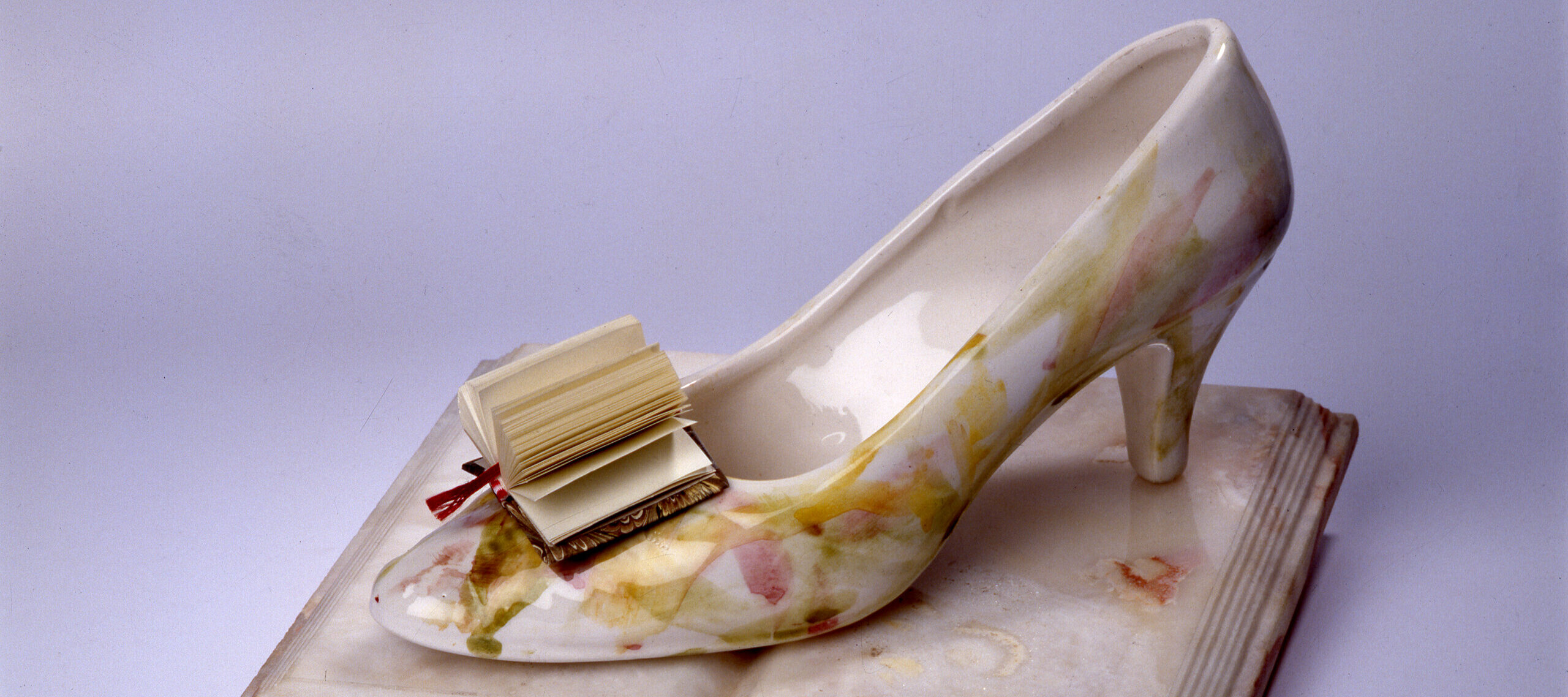 A woman's glass slipper rests on the open pages of a porcelain book. In place of a bow on the toe of the slipper is a paper book with pages fanned out.
