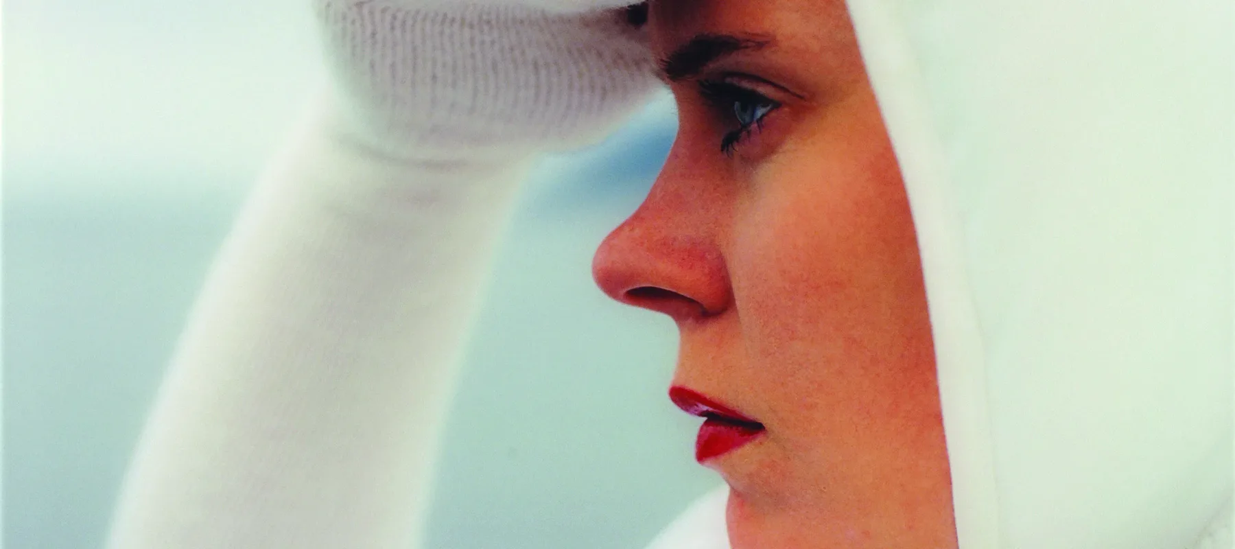 Square close-up of a light-skinned woman in profile looking to the left, seen from the shoulders up. She wears all white, including a bonnet and knit sleeves that become gloves covering her three middle fingers, and she holds her right hand up as if shielding her eyes from the sun.