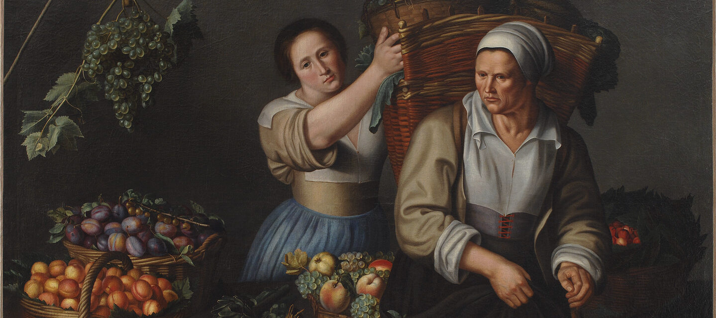 Two women are surrounded by baskets of grapes and other produce. They are both light-skinned and wearing old-fashioned attire with head wraps and full skirts, stand on either side of a table, as the younger one places a bin of grapes into a large basket on the other’s back.