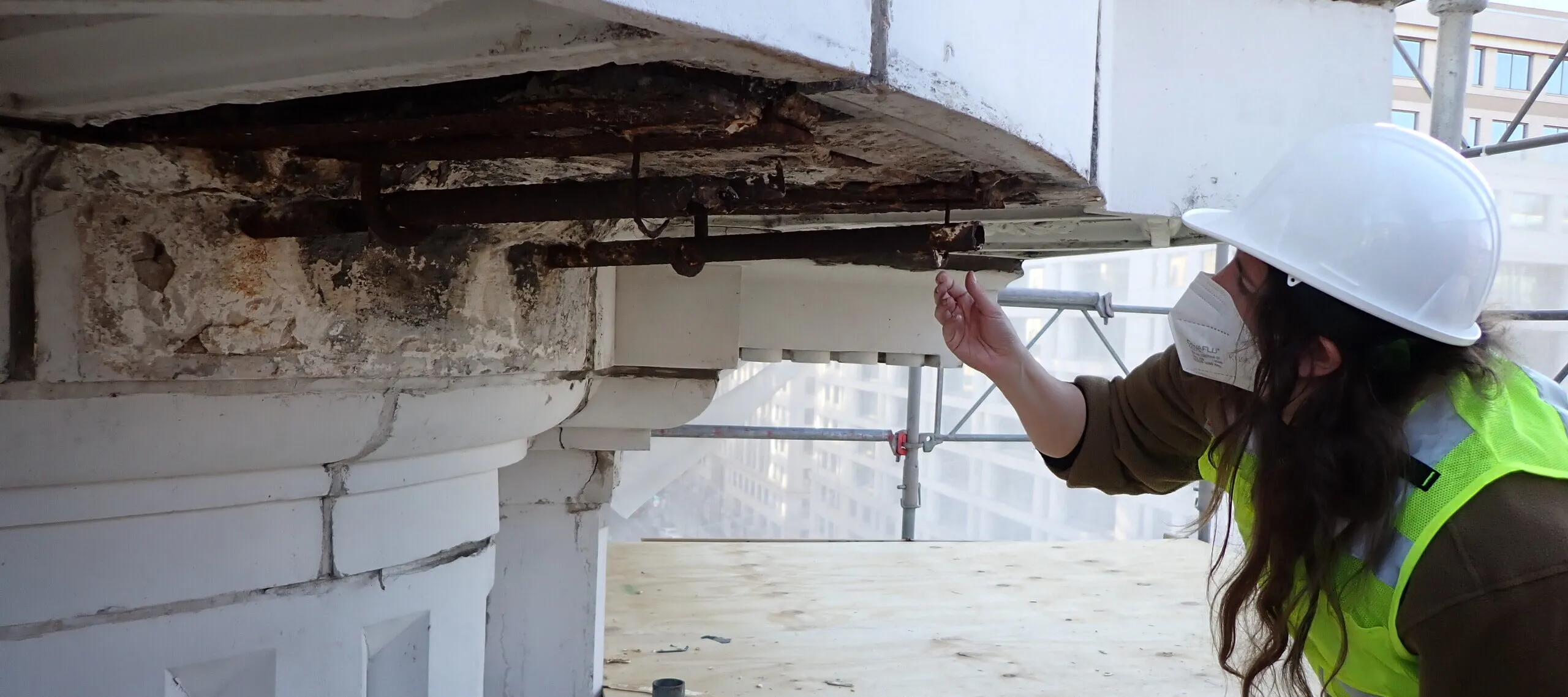 A light-skinned woman wearing a yellow safety vest, a white hard hat, and a white face mask inspects the cornice on the roof of NMWA.