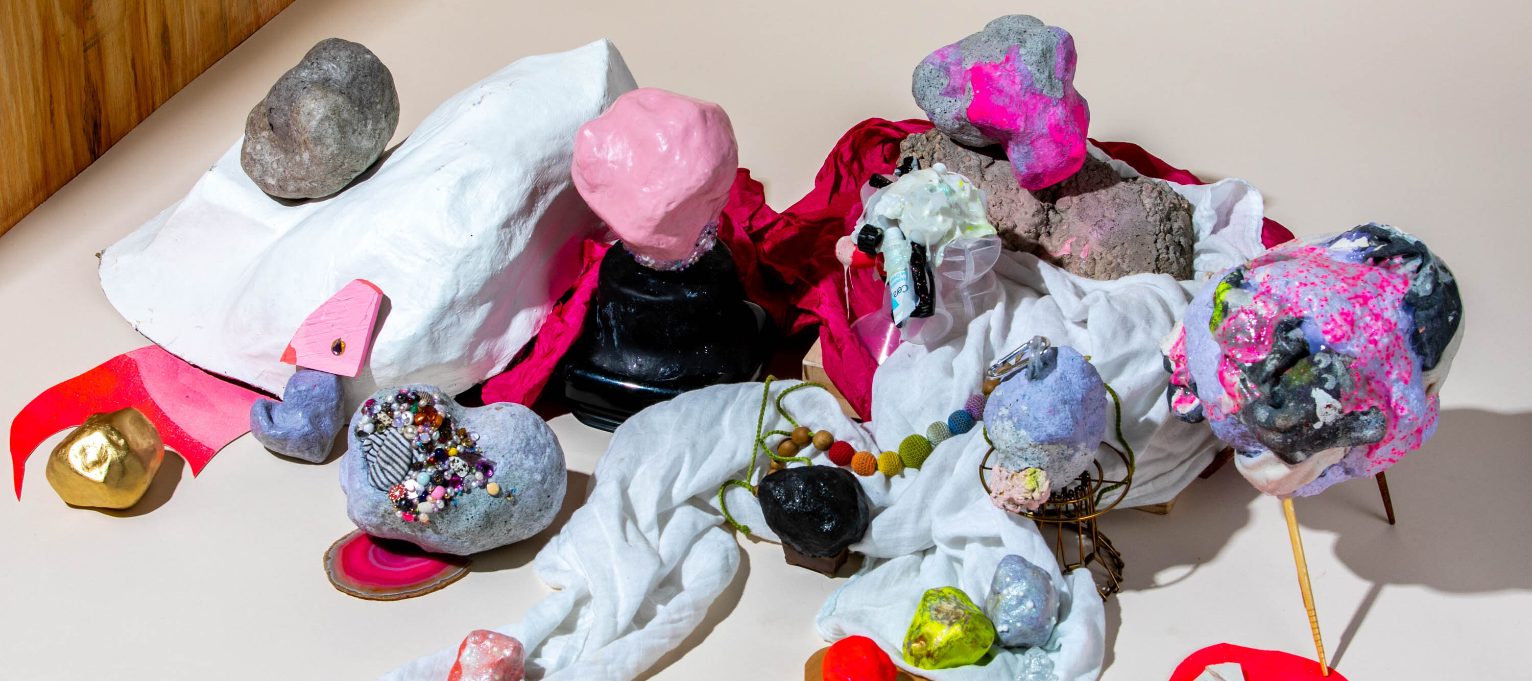 Header image: An assemblage of a variety of objects with different textures such as stones, textiles, beads, wood, and paper. The objects are painted mainly in red and pink colors and stacked on top of each other, creating a chaotic, colorful scene.