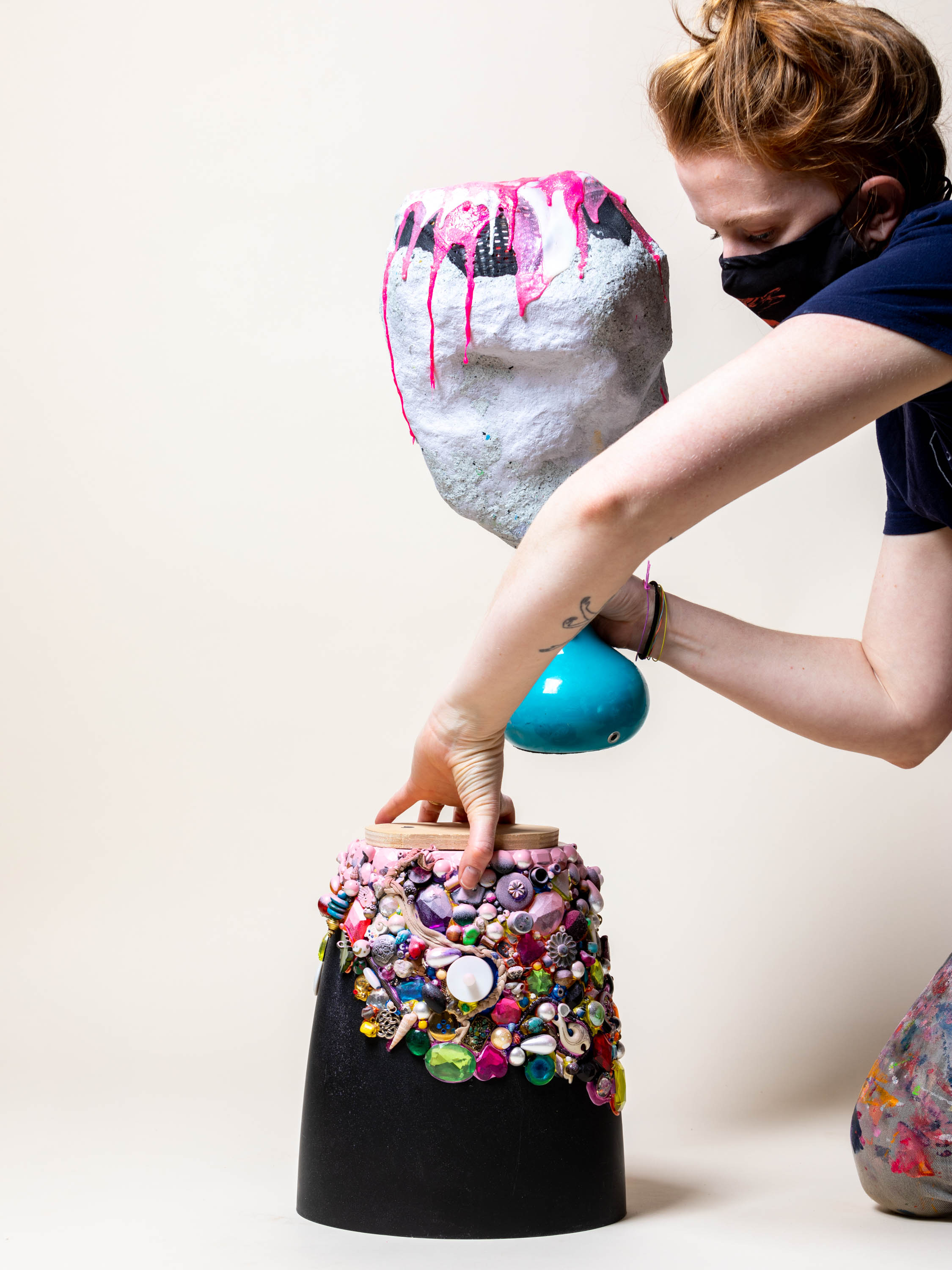 A woman with red hair in a bun and a light skin tone holds a colorful sculpture in her right hand and holds another one that sits on the floor. The object on the floor consists of a black pot covered in little objects such as shells, beads, and pearls.