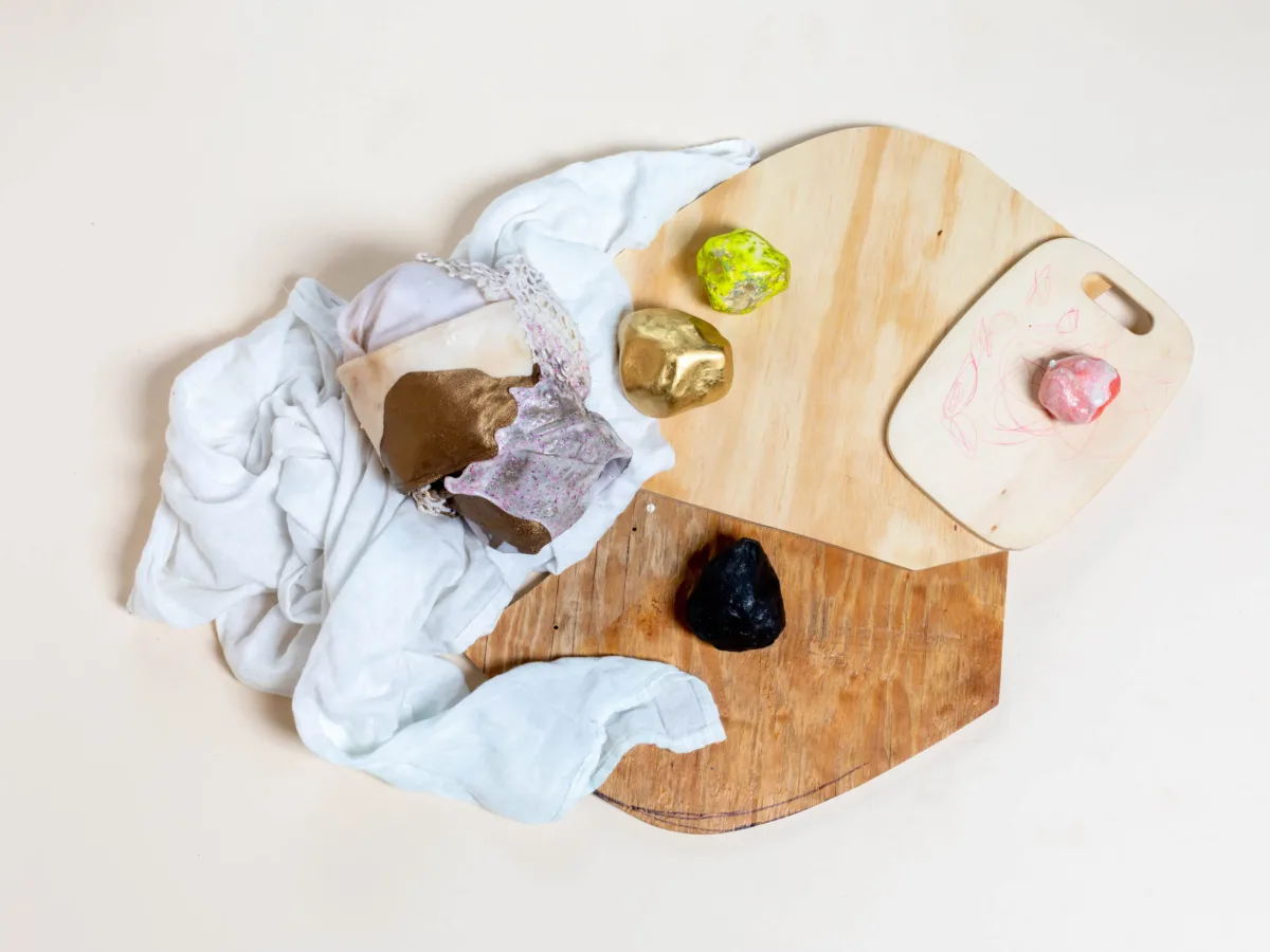 An assemblage consisting of wooden pieces, a cloth, and painted stones. The stones are painted in black, golden, and yellow. The objects are arranged on top of each other and the textures juxtapose each other.