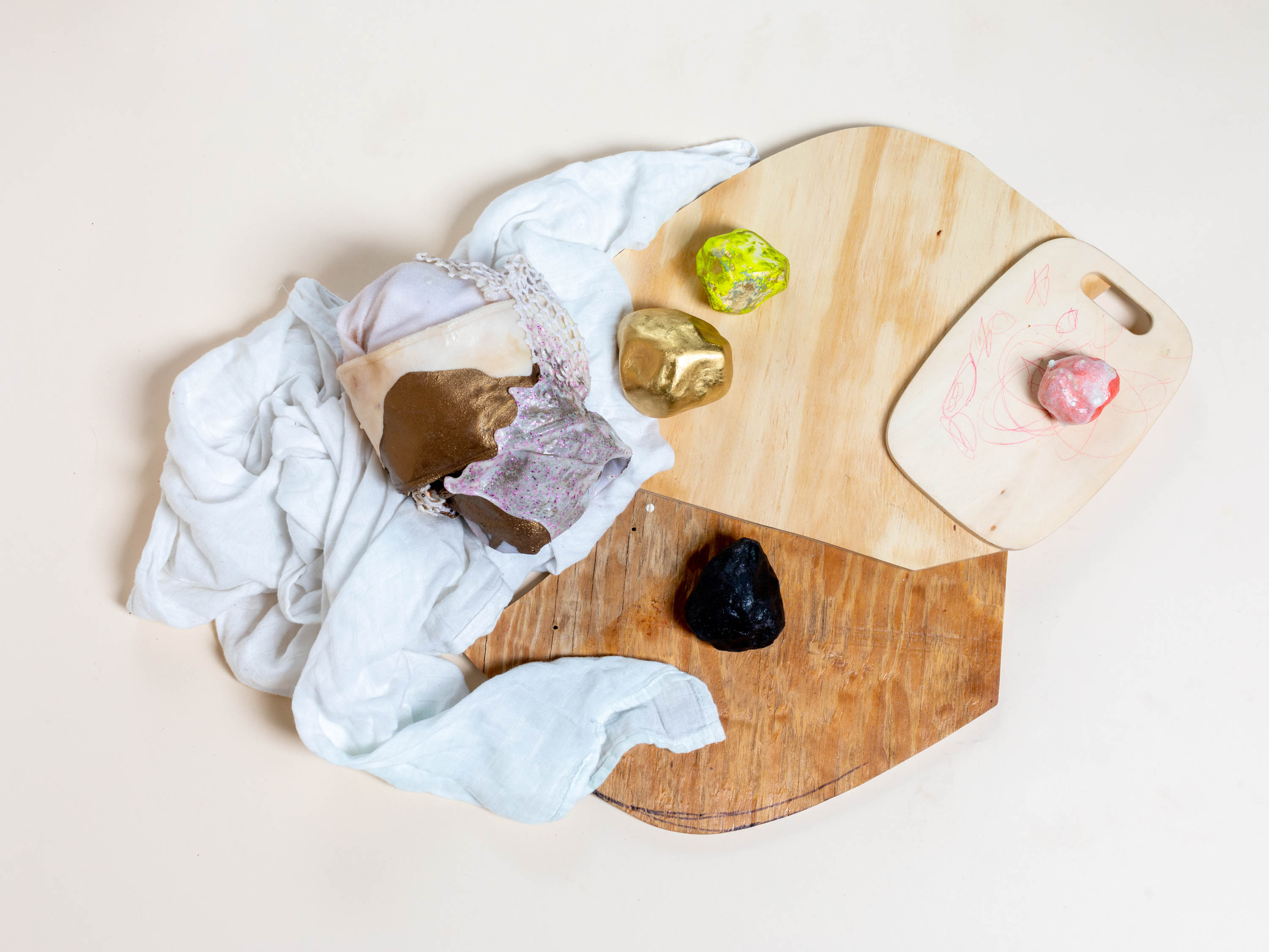 An assemblage consisting of wooden pieces, a cloth, and painted stones. The stones are painted in black, golden, and yellow. The objects are arranged on top of each other and the textures juxtapose each other.