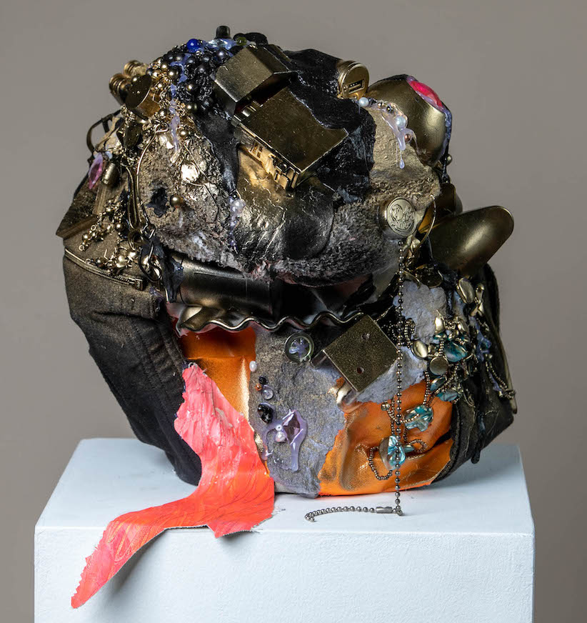 A round sculpture with many different objects attached to it. A variety of textures creates an interesting surface. Paper, metal, beads, and textiles in black, orange and gold are glued onto it.