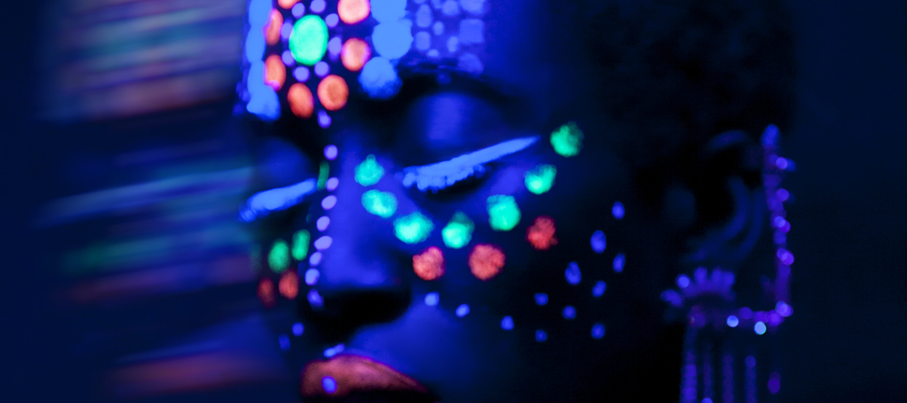 A dark-skinned woman stands in a dark space illuminated by a black light. Her face is painted with green, orange, and white dots and orange lipstick that all glow in the black light. She wears a beaded top and an earring with strands of beads that run from her earlobe to the top of her ear.