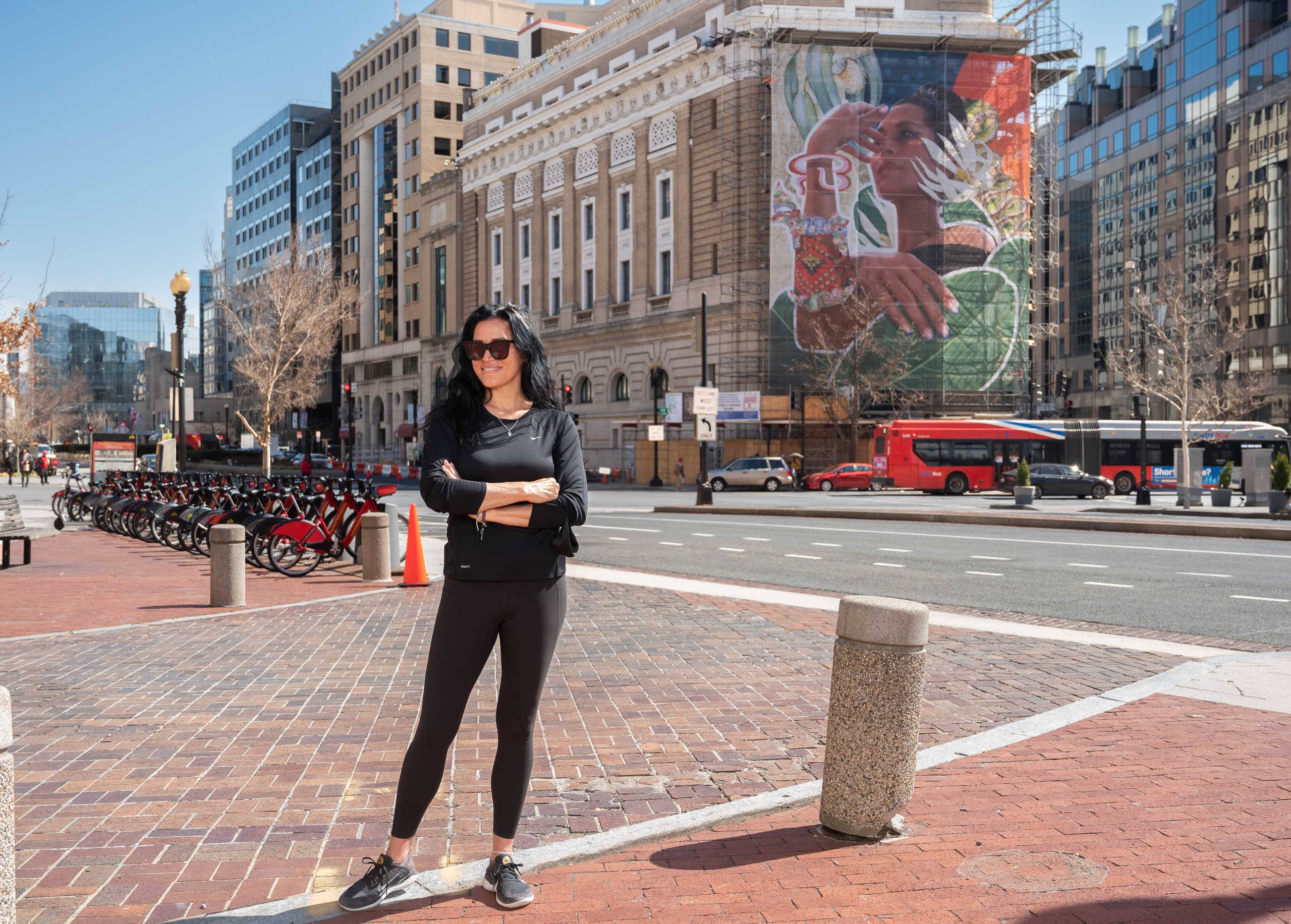 Cita Sadeli grins and stands with her arms crossed. She is a medium-light skinned adult woman with straight, black hair, wearing all black and sunglasses. Behind her, her colorful, four-story tall portrait of a medium-skinned adult woman surrounded by greenery hangs from a building.