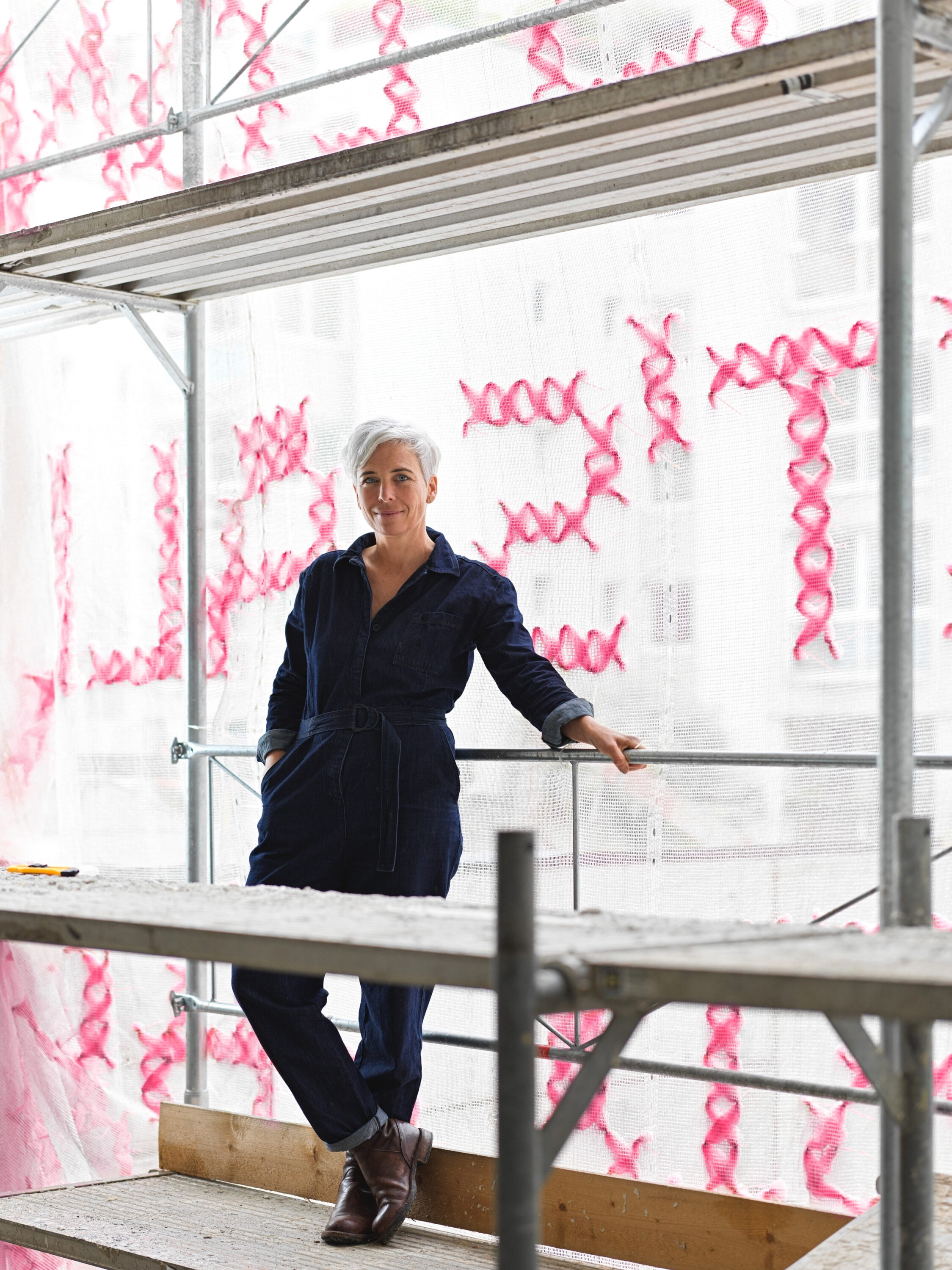 A light-skinned woman with short white hair stands on a scaffolding platform. Behind her, the scaffolding is covered in white mesh scrim stitched with bright pink letters.