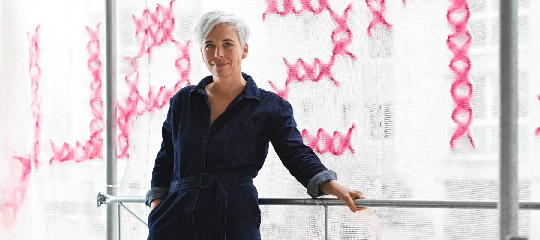 A light-skinned woman with short white hair stands on a scaffolding platform. Behind her, the scaffolding is covered in white mesh scrim stitched with bright pink letters.