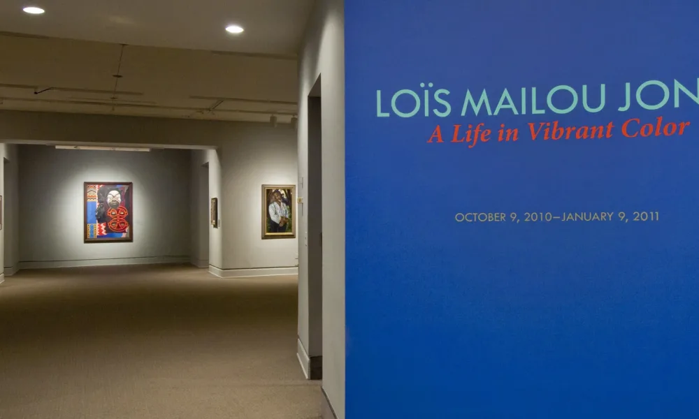 Intervior view of the museum galleries features a vibrant blue wall with the text 'Lois Mailou Jones: A Life in Vibrant Color'. Multiple artworks are displayed in the background.