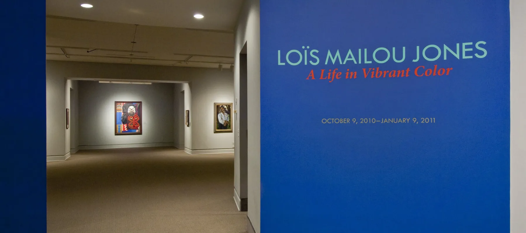 Intervior view of the museum galleries features a vibrant blue wall with the text 'Lois Mailou Jones: A Life in Vibrant Color'. Multiple artworks are displayed in the background.