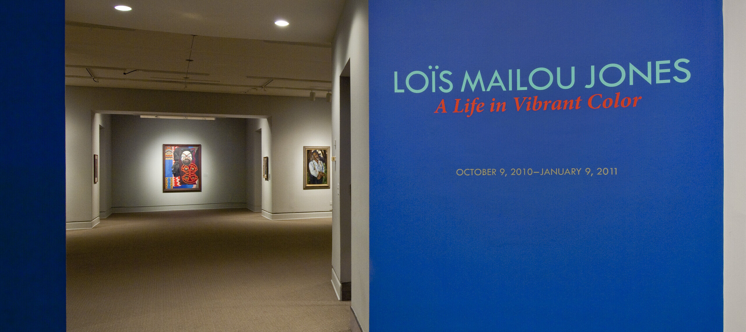 Intervior view of the museum galleries features a vibrant blue wall with the text 'Lois Mailou Jones: A Life in Vibrant Color'. Multiple artworks are displayed in the background.