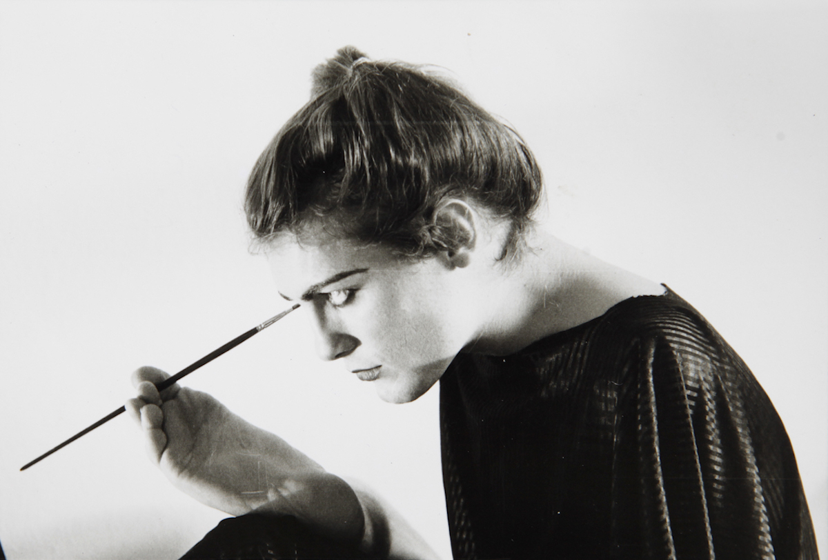 A black-and-white photograph of a woman with a light skin tone who is holding a paintbrush with her foot. She wears a black dress and her hair in a ponytail.