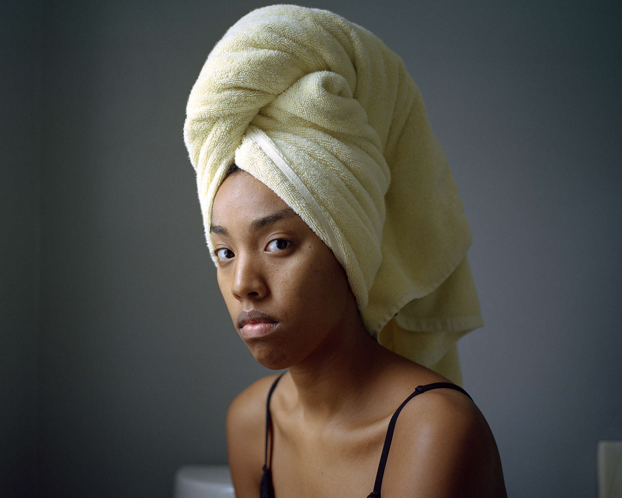 A woman with a medium-dark skin tone wears a yellow towel wrapped around her hair. She is looking directly at the camera with a questioning glance, sitting before an empty dark grey wall. 