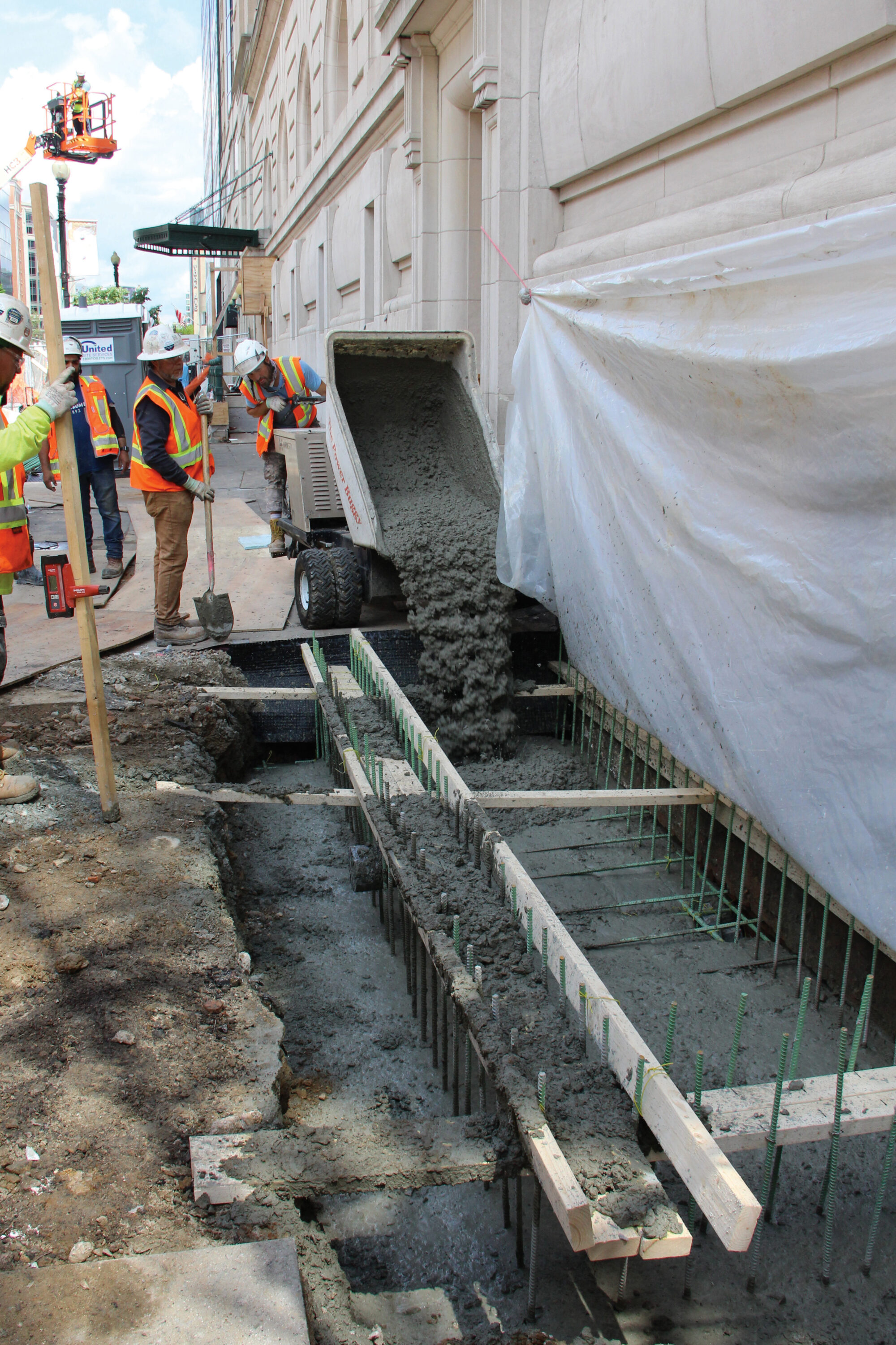 Members of a construction team pour concrete to fill a trench in the sidewalk outside of NMWA’s historic stone building. The dark gray concrete flows from the upturned back of a small truck as several people in orange-and-yellow vests and white protective helmets look on.