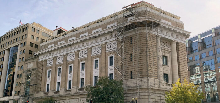 The National Museum of Women in the Arts’ historic Renaissance Revival building is pictured on a sunny day in downtown Washington, D.C. Scaffolding is set up on the front corner of the wedge-shaped structure, from top to bottom. On the sidewalk below, there is a covered walkway, panels of wood covering the doors and windows, and orange-and-white construction barriers.