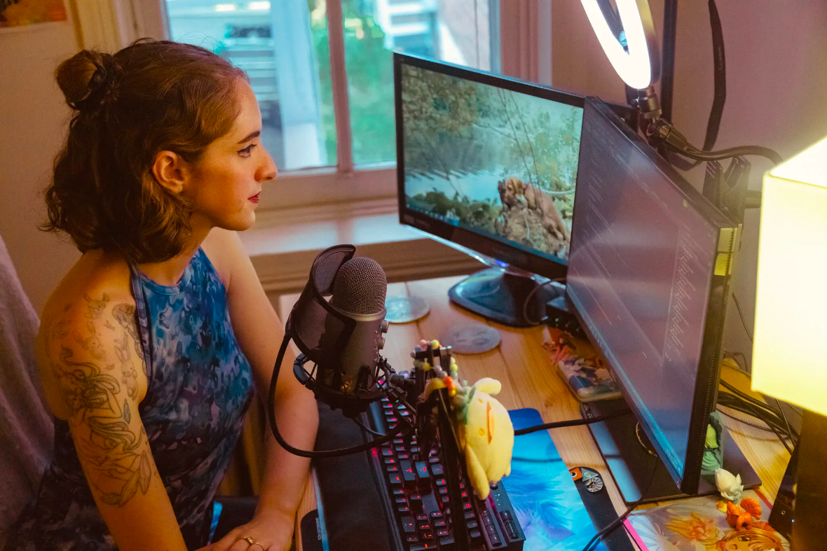 A young woman with a light skin tone, short red hair, and a sleeve tattoo sits in her room. She looks at two computer screens in front of her; one of them depicts a coding application.