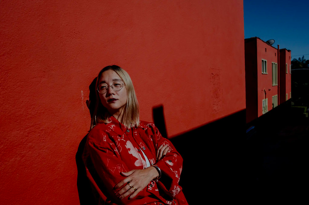 A portrait of an Asian American woman leaning against a red wall. She is wearing a red jacket with abstract forms, black lipstick, and glasses. 