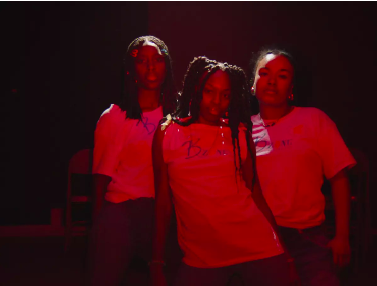 A film still of three young women with a dark skin tone in white T-shirts and with long braided hair. The women look directly into the camera and are bathed in a red and purple light.