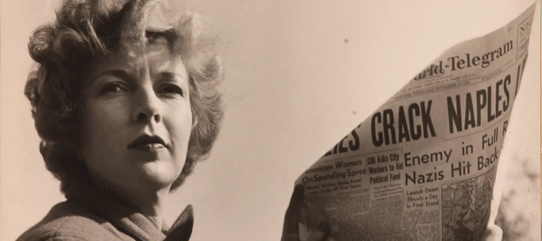 A black-and-white photograph of a light-skinned adult woman holding a newspaper with news about World War II. She wears a coat and her short, curly hair is caught in the wind.